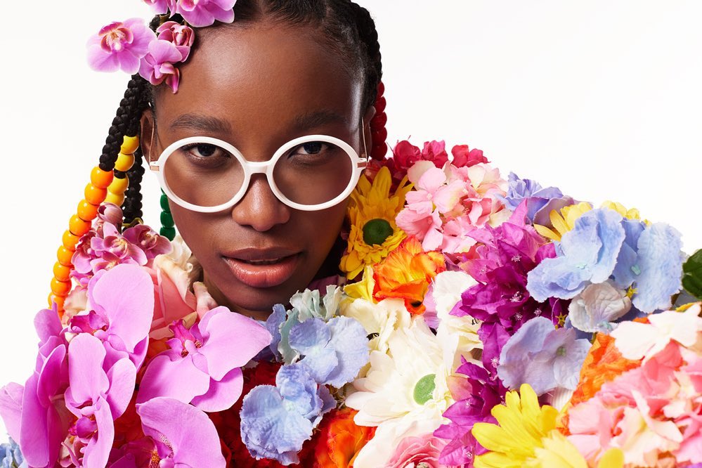 Eye-level portrait of a Black woman with braids, wearing glasses, surrounded by colorful flowers against a bright white background.