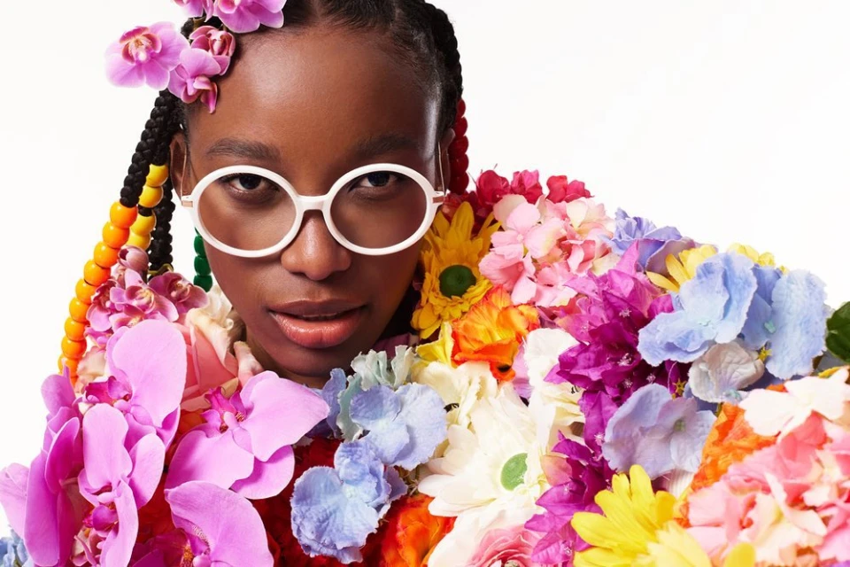 Eye-level portrait of a Black woman with braids, wearing glasses, surrounded by colorful flowers against a bright white background.