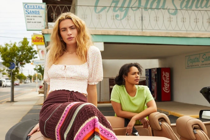 Two women pose on a car in front of the Crystal Sands motel. The woman in front is wearing a floral top and knitted skirt.
