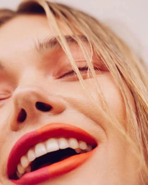 Close up of a smiling adult woman with blonde hair and orange lipstick, showing white teeth in an indoor setting.
