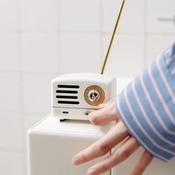 Close-up of a hand operating a small white vintage radio with a gold antenna on top of a white cube in a white-tiled room.