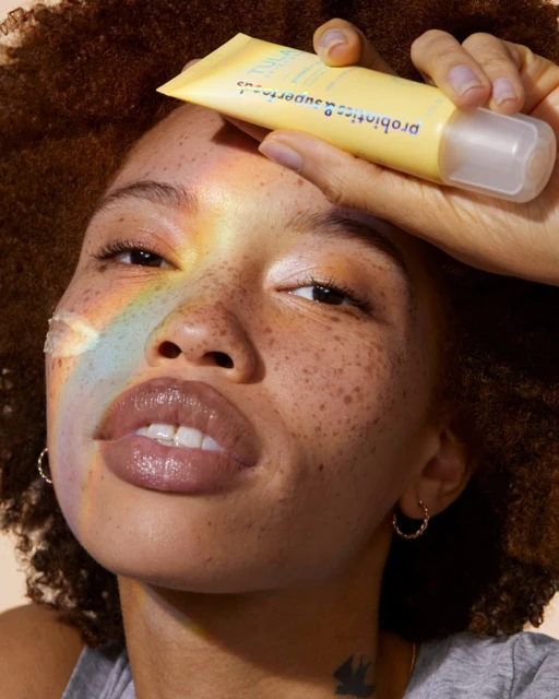 Close-up beauty shot of a woman with freckles holding a yellow tube of Tula Probiotic skincare product up to her forehead.