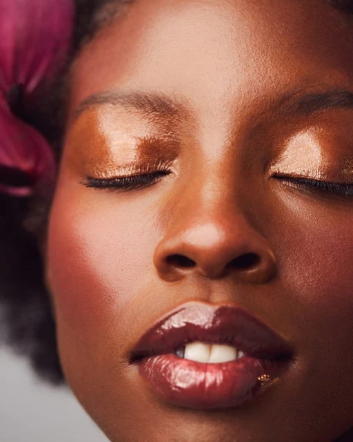 Close-up portrait of an African-American female with glossy brown lips and metallic bronze eyeshadow, accented by a purple flower.
