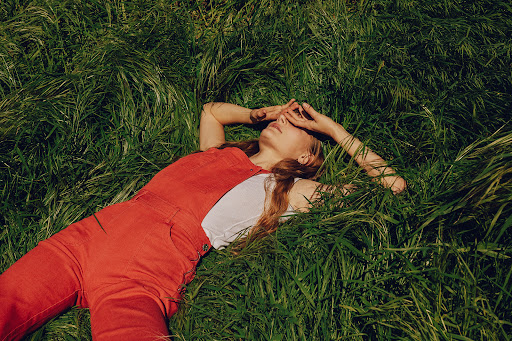 Young woman in red overalls lying in green grass, covering her eyes with her hands. Sunny day, outdoor setting. Fashion and lifestyle.
