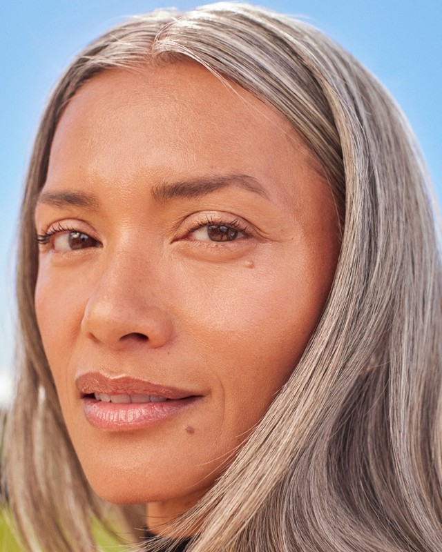 Close-up portrait of an adult woman with graying hair against a blurred background of sky and green, highlighting her natural beauty.
