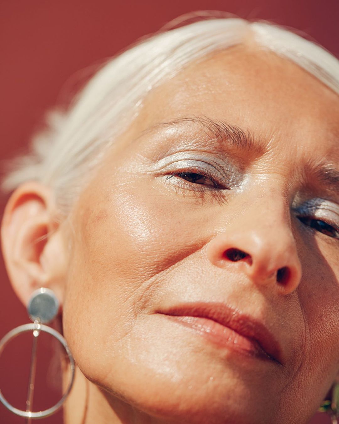 Close-up portrait of an older adult woman with silver eyeshadow and hoop earrings against a solid dark-red background.