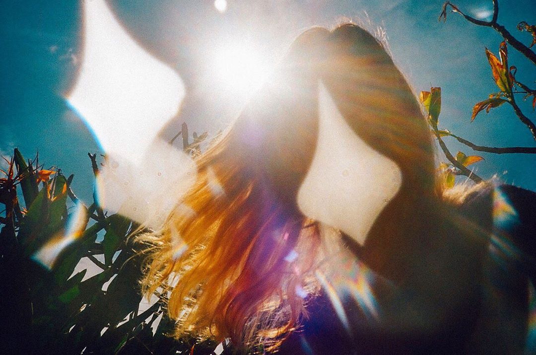 Low angle view of a woman with long wavy hair, sunlight flares, and tropical plants and sky in the background. 