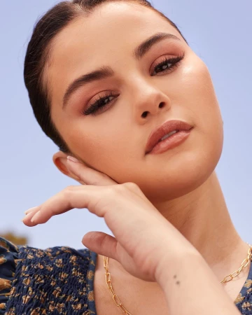 Close-up portrait of Selena Gomez with warm neutral makeup holding her hand near her face against a clear blue sky