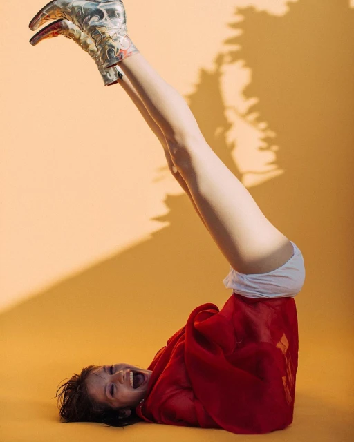 A cheerful woman with legs up wearing cowboy boots lies on an orange background, covered by a red blanket.