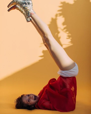 A cheerful woman with legs up wearing cowboy boots lies on an orange background, covered by a red blanket.