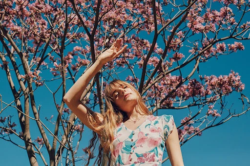 Young woman in a floral dress poses with her arm raised in front of a flowering tree against a blue sky in outdoor setting.