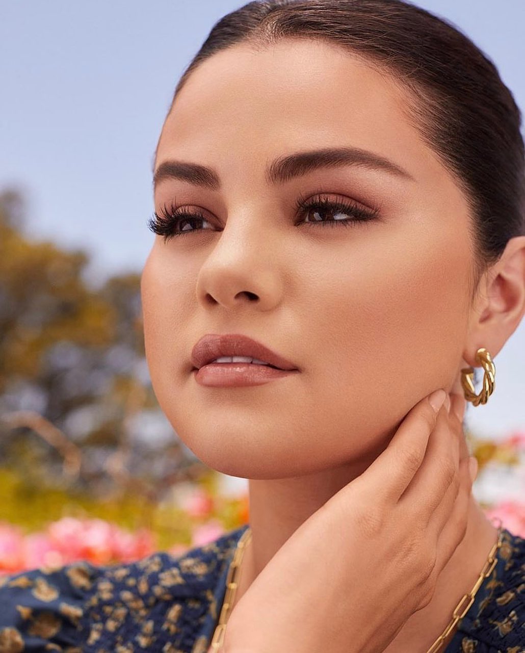 Close-up beauty shot of Selena Gomez wearing gold earrings and a patterned top, posing outdoors with a soft, blurred background.