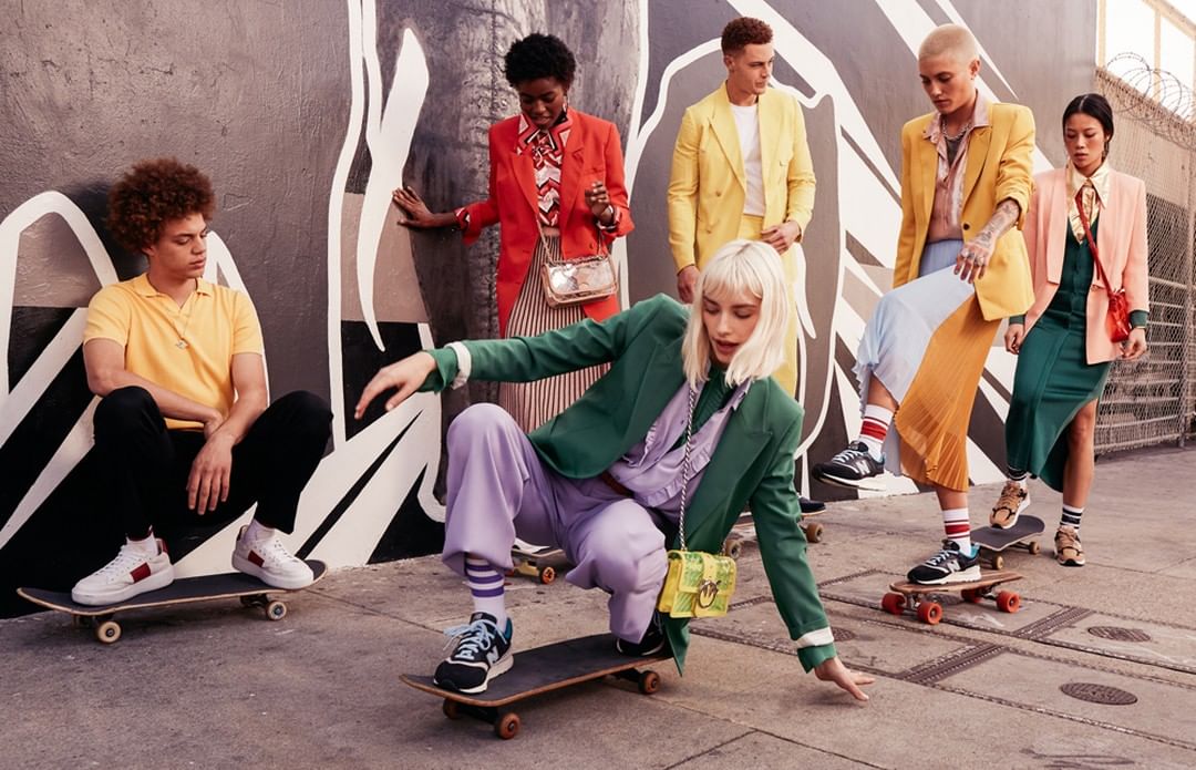 Group of stylish young adult models on skateboards in an urban setting, wearing bright colored blazers and trendy outfits.