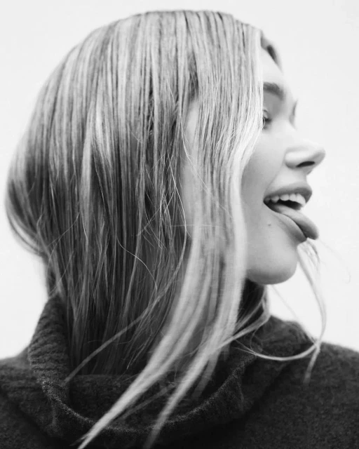 Close-up black and white portrait of a young adult woman sticking her tongue out with blonde hair against a plain background.