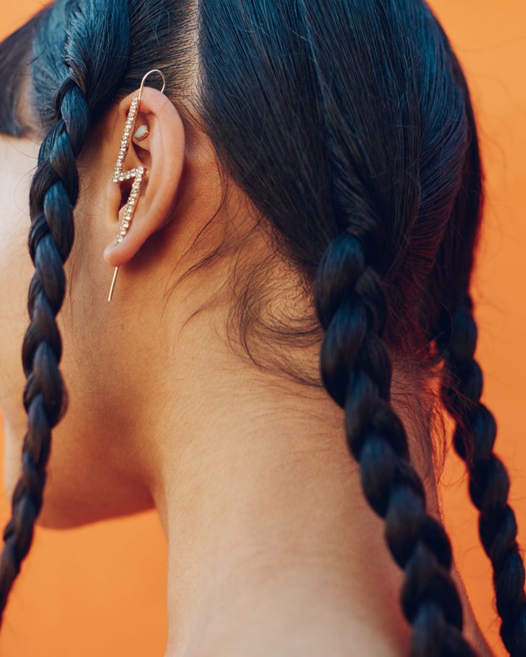 Close-up of a woman's head with braided black hair, featuring a rhinestone lightning bolt earring on an orange background.