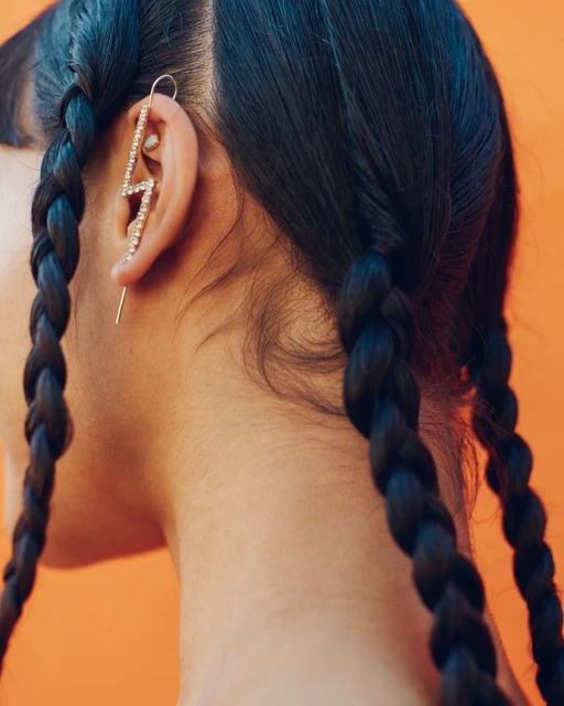 Close-up of a woman's head with braided black hair, featuring a rhinestone lightning bolt earring on an orange background.