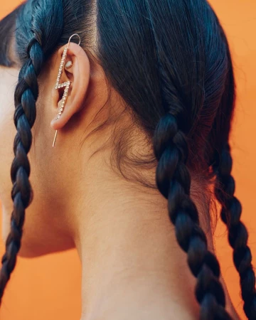 Close-up of a woman's head with braided black hair, featuring a rhinestone lightning bolt earring on an orange background.