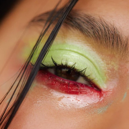 Extreme close-up of an eye with vibrant green eyeshadow and red liner, featuring dark strands of hair falling across the frame.