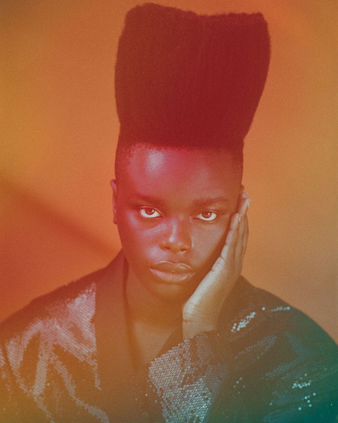 Portrait of a non-binary person with a striking high-top fade haircut, posing in front of a gradient orange and red background.