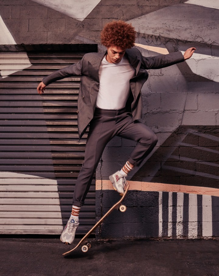 A young man with curly red hair jumps mid-air with a skateboard, wearing a grey suit jacket, white t-shirt, and patterned socks.