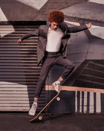 A young man with curly red hair jumps mid-air with a skateboard, wearing a grey suit jacket, white t-shirt, and patterned socks.