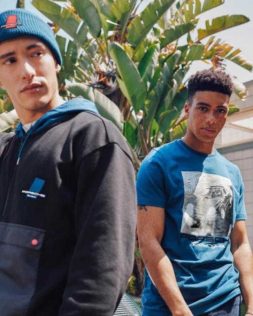 Two men wearing Porsche branded apparel stand outdoors against a backdrop of green foliage and neutral buildings. Automotive fashion.