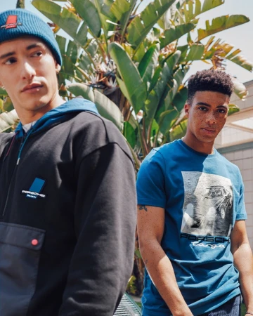 Two men wearing Porsche branded apparel stand outdoors against a backdrop of green foliage and neutral buildings. Automotive fashion.