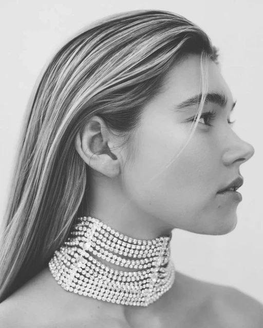 Black and white portrait of a young woman with long blonde hair wearing a multi-strand pearl choker against a plain white background.
