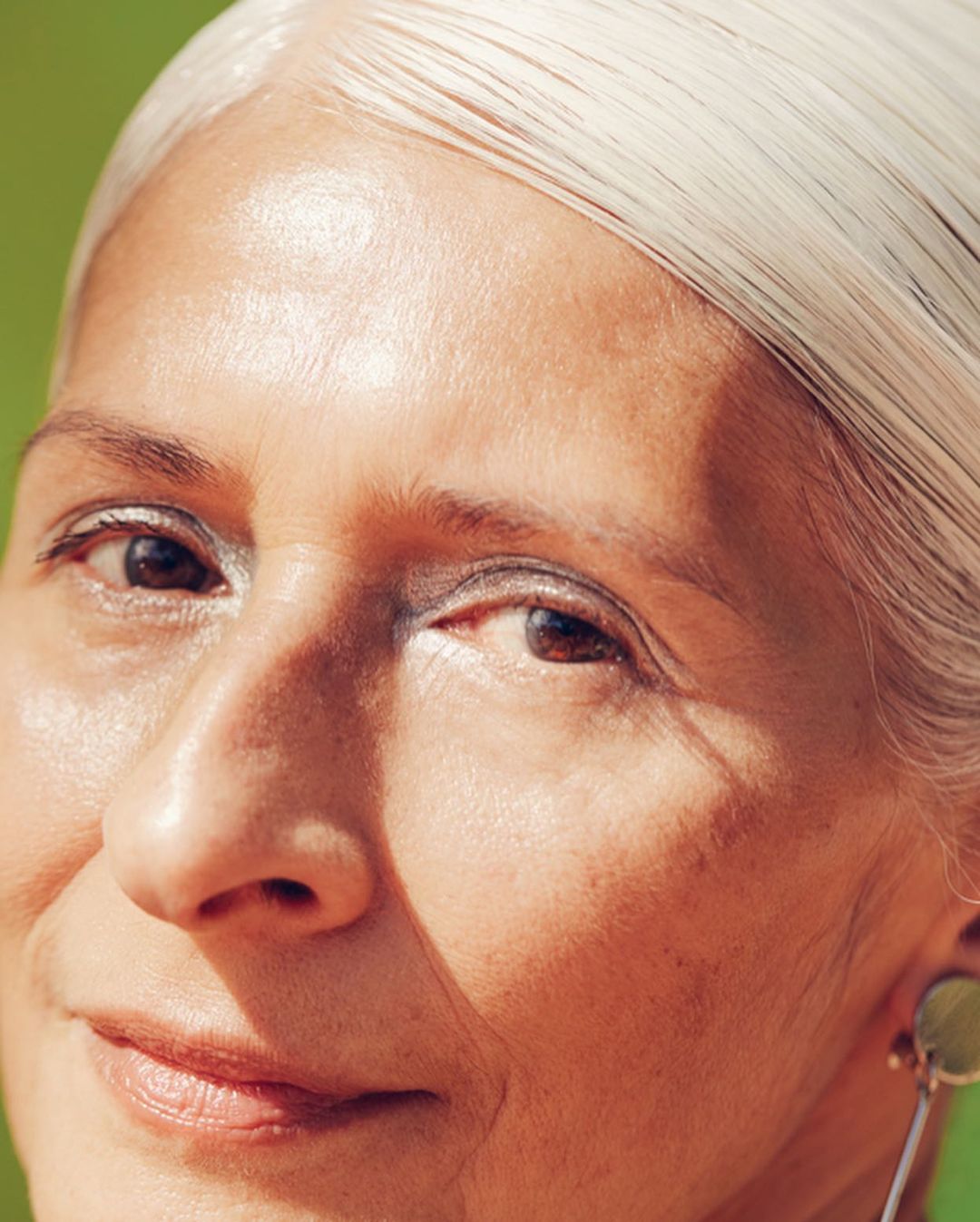 Close-up portrait of an older woman with straight silver hair and natural skin texture against a green background