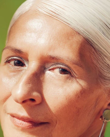 Close-up portrait of an older woman with straight silver hair and natural skin texture against a green background