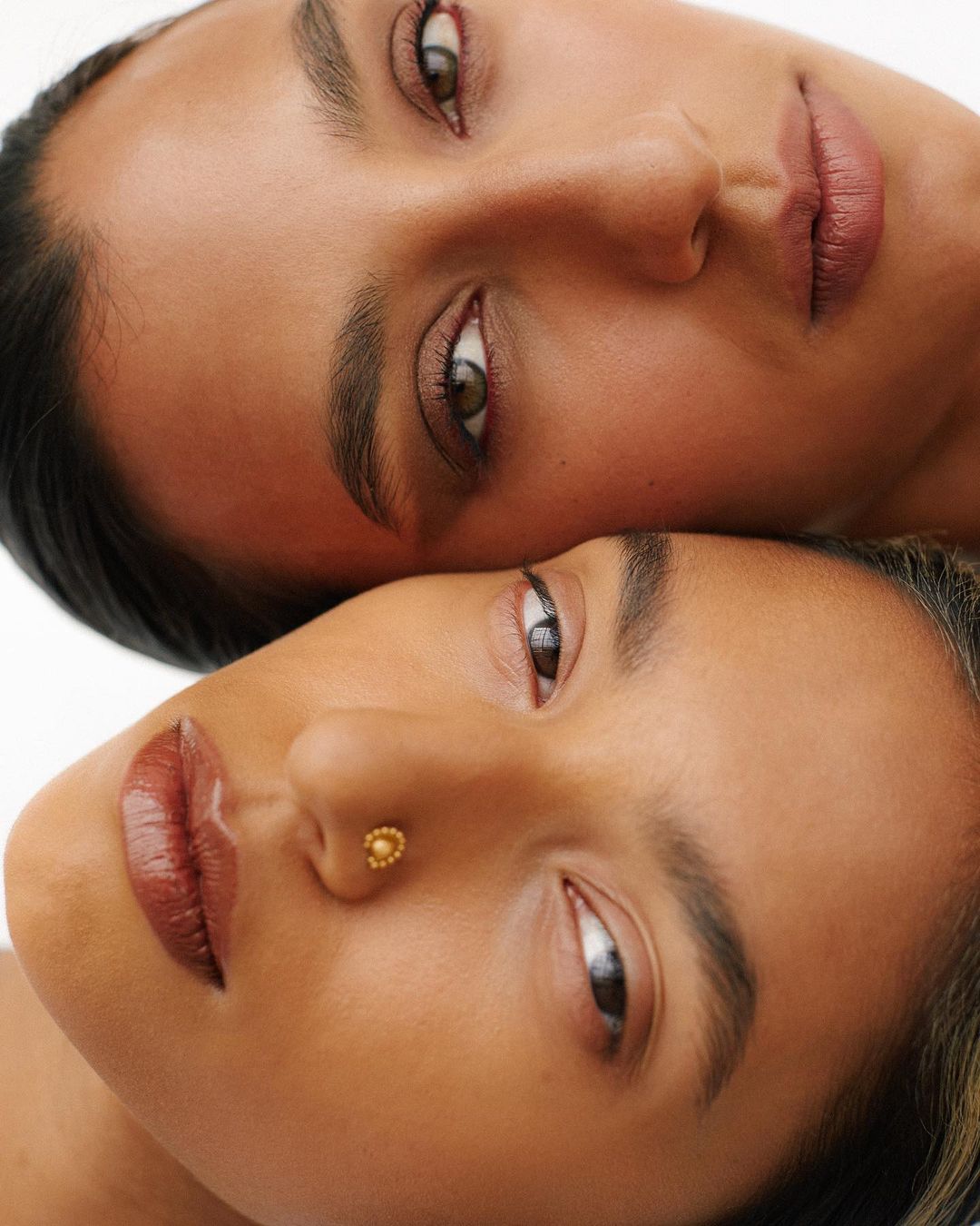 Close-up portrait of two young adult women with bronze skin tones indoors in a beauty or skincare context with soft lighting.