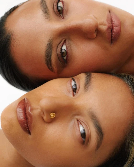 Close-up portrait of two young adult women with bronze skin tones indoors in a beauty or skincare context with soft lighting.