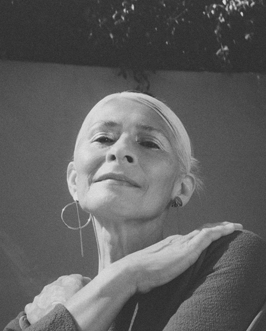 Black and white portrait of an older adult woman with short white hair wearing modern earrings, with foliage visible above.
