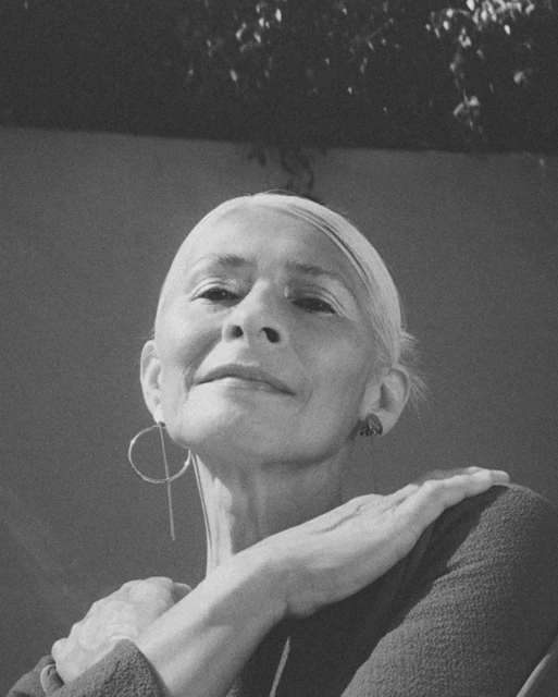 Black and white portrait of an older adult woman with short white hair wearing modern earrings, with foliage visible above.