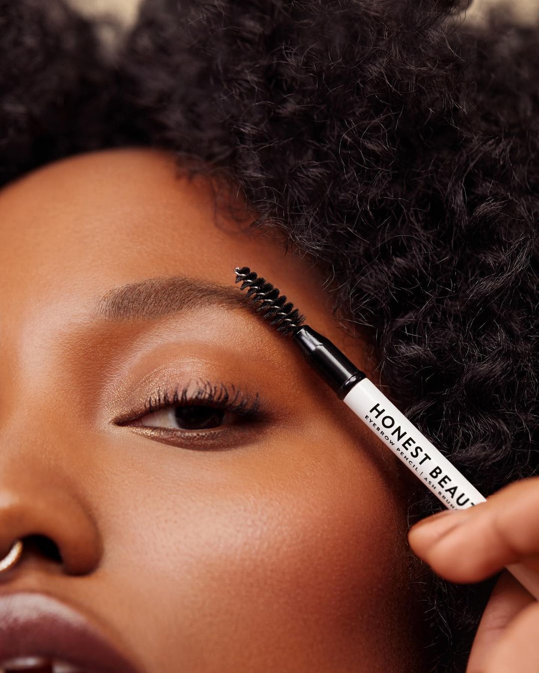 Woman applies eyebrow pencil to eyebrow for a beauty shot. Close-up of a female face with dark hair and skin. Beauty product shown.