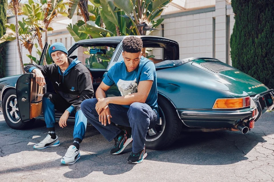 Two men wearing Puma apparel pose with a dark green classic Porsche car. Outdoor setting with palm trees and a building.