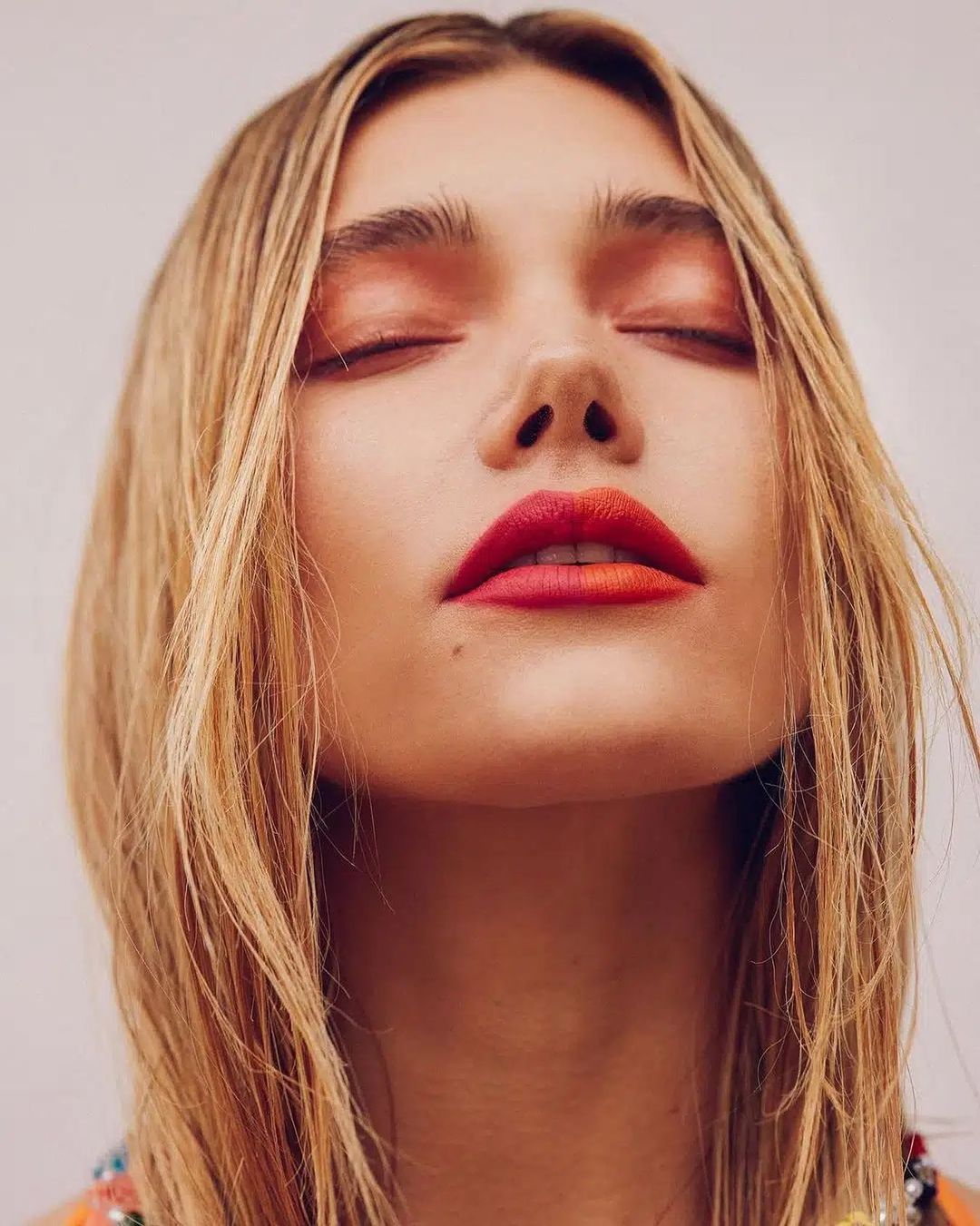 Close-up portrait of a blonde woman with eyes closed, wearing bold orange and red gradient lip color, and light orange eyeshadow.