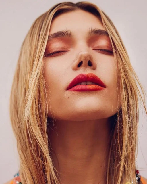 Close-up portrait of a blonde woman with eyes closed, wearing bold orange and red gradient lip color, and light orange eyeshadow.