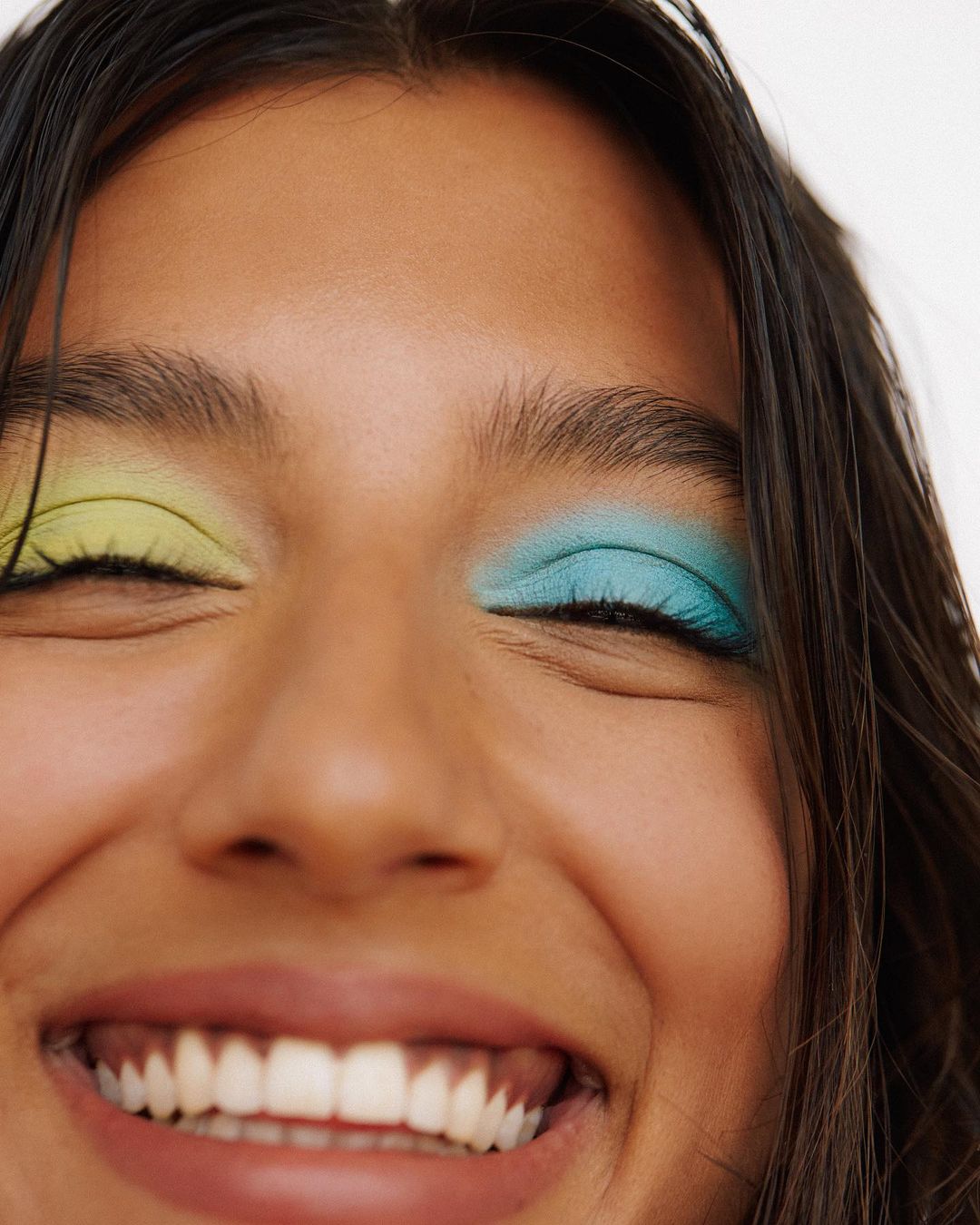 Close-up of a smiling woman with vibrant yellow and teal eyeshadow, showcasing a joyful and colorful beauty aesthetic.