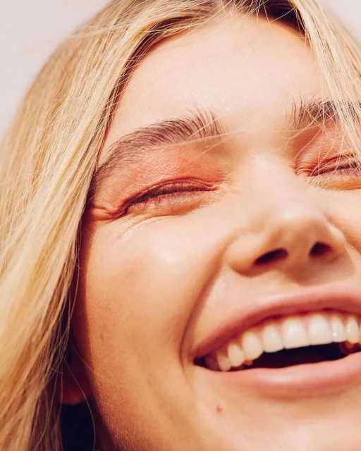 Close-up portrait of a smiling young woman with blonde hair and light skin, showcasing pink eyeshadow against a light background.