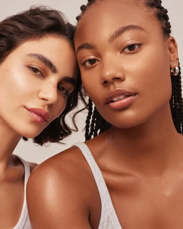 Close-up beauty portrait of two young women with glowing skin wearing white tank tops against a plain background by Kayla Varley