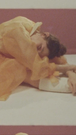 Adult female laying down in a yellow robe on a white surface with arms crossed under her face, the backdrop is mauve.