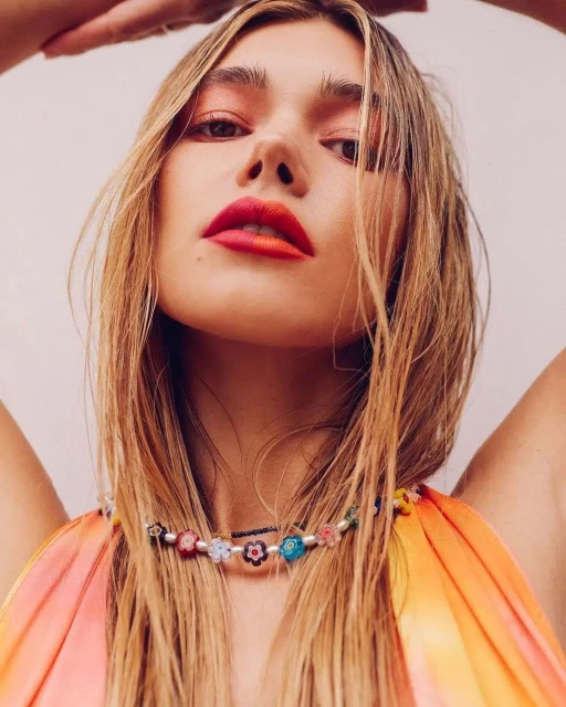 Close-up shot of a woman with blonde hair and vibrant makeup, wearing a colorful beaded necklace against a light background.