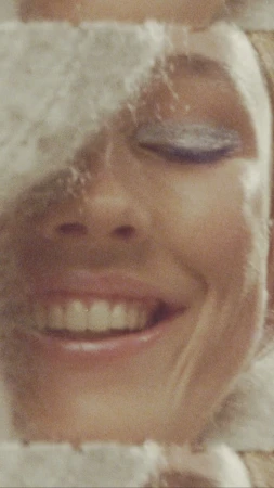 Close-up portrait of a smiling adult female with soft makeup and a white, fluffy head covering, showcasing a beauty and fashion aesthetic.