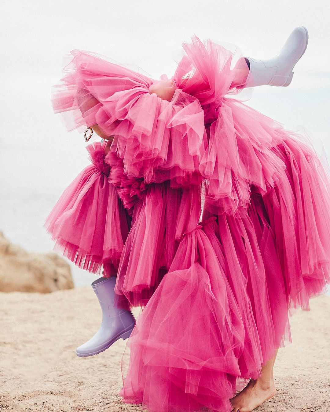 Fashion shot of a person wearing a pink layered tulle dress with boots while posing on a sandy surface with a pale sky background.