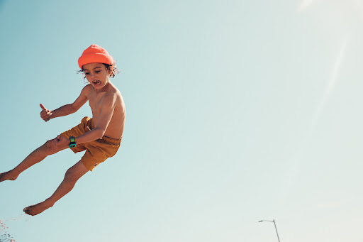 A young child with curly hair and an orange beanie jumps against a clear blue sky, wearing yellow shorts and a blue and green watch.