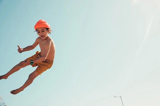 A young child with curly hair and an orange beanie jumps against a clear blue sky, wearing yellow shorts and a blue and green watch.