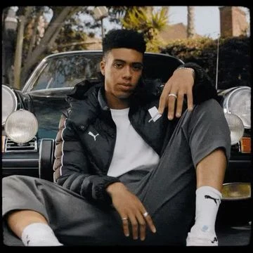 A young Black man wearing a black puffer jacket and grey trousers seated in front of a classic car.