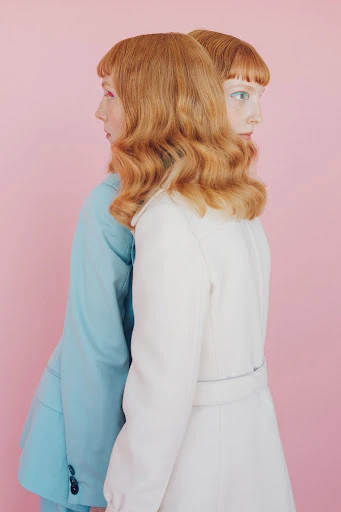 Two women with red hair stand back to back in a studio setting. One wears a light blue suit and the other a light beige coat.