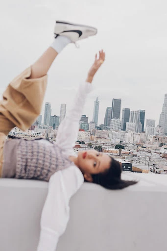 A woman lies on her back with legs up in the air wearing sneakers, against a cityscape backdrop on an overcast day, medium outdoor shot.